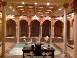 The image shows an ornate indoor spa or bathhouse with a luxurious design. The room features a central hot tub or small pool surrounded by intricately carved columns and arches. The ceiling is decorated with detailed patterns and recessed lighting. In the foreground, there is a table with various spa items, including towels, candles, and a vase with flowers. The overall ambiance is elegant and serene.