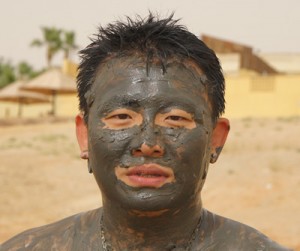 A person with short, dark hair is shown with their face covered in mud. The background appears to be an outdoor setting with some buildings and palm trees visible in the distance. The person is looking directly at the camera.