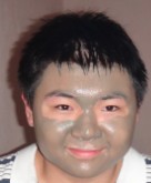 A person with short black hair is smiling at the camera. Their face is covered with a gray facial mask. They are wearing a white shirt with black stripes. The background is plain and out of focus.