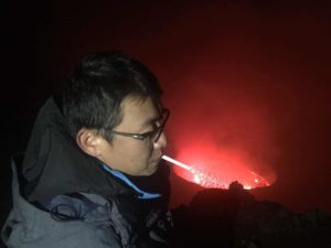 A person wearing glasses and a dark jacket is standing near the edge of a glowing red crater, possibly a volcano, at night. The red glow from the crater illuminates the surrounding area, creating a dramatic and intense atmosphere.