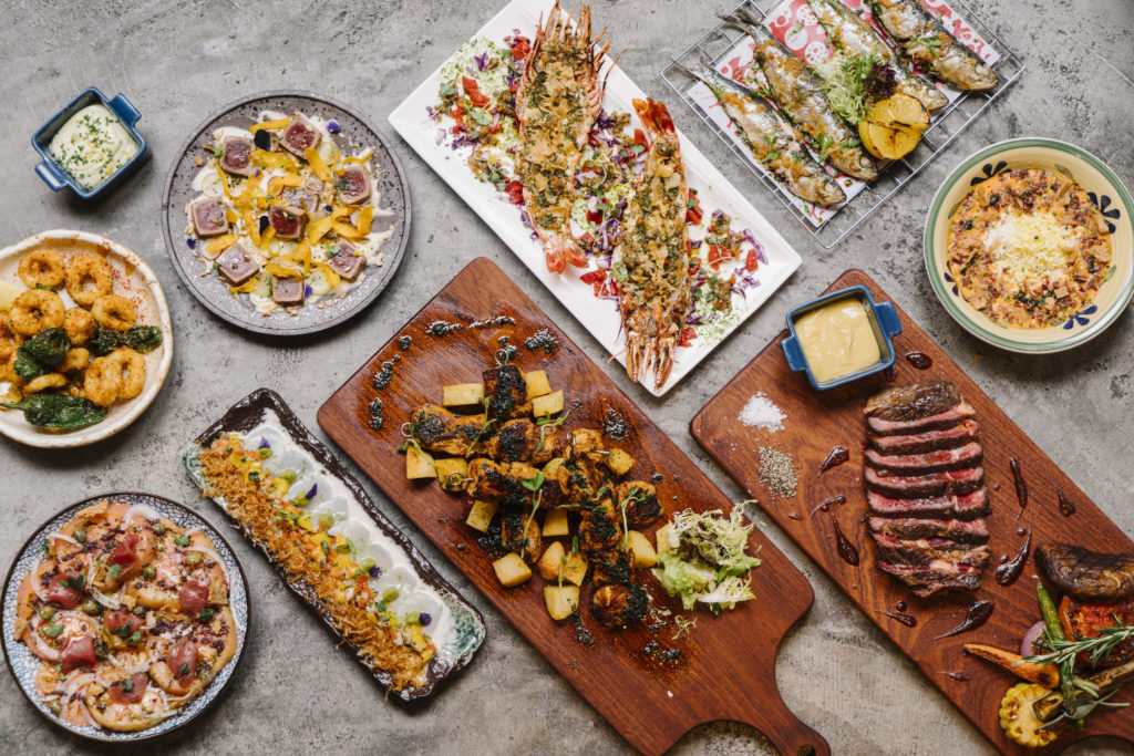 A variety of gourmet dishes are arranged on a table. The spread includes grilled prawns on a rectangular white plate, grilled fish on a wire rack with lemon slices, a bowl of risotto, a wooden board with sliced steak and condiments, a plate of seared tuna with garnishes, a dish of fried calamari, a long plate with a seafood dish topped with herbs, and a wooden board with roasted vegetables and potatoes. There is also a small bowl of sauce and a bowl of creamy dip. The table surface is a textured gray.