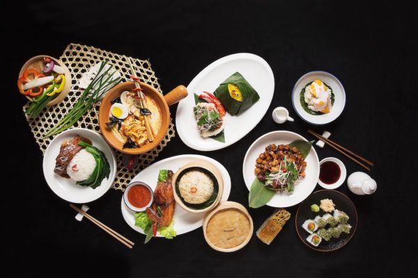 Alt text: A variety of Asian dishes are arranged on a black surface. The dishes include rice, noodles, vegetables, and various meats, presented in bowls and plates. There are also dipping sauces, a bamboo steamer with buns, and a small bowl of soup. Chopsticks and a teapot with cups are also visible. The food is garnished with herbs and vegetables, creating a colorful and appetizing display.