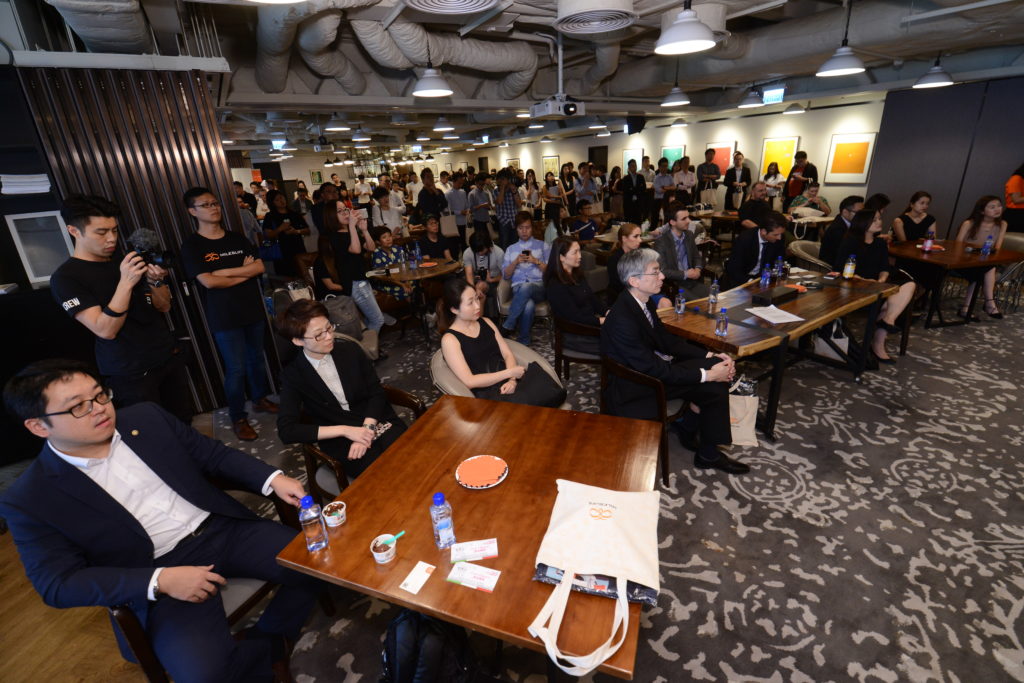The image shows a large group of people gathered in a modern, well-lit room with industrial-style ceiling fixtures. The attendees are seated at tables or standing, facing towards the front of the room, suggesting they are listening to a presentation or speaker. Some people are taking notes, while others are using their phones or cameras. The tables have water bottles, papers, and other small items on them. The room has a patterned carpet and artwork displayed on the walls. The atmosphere appears to be professional and attentive.
