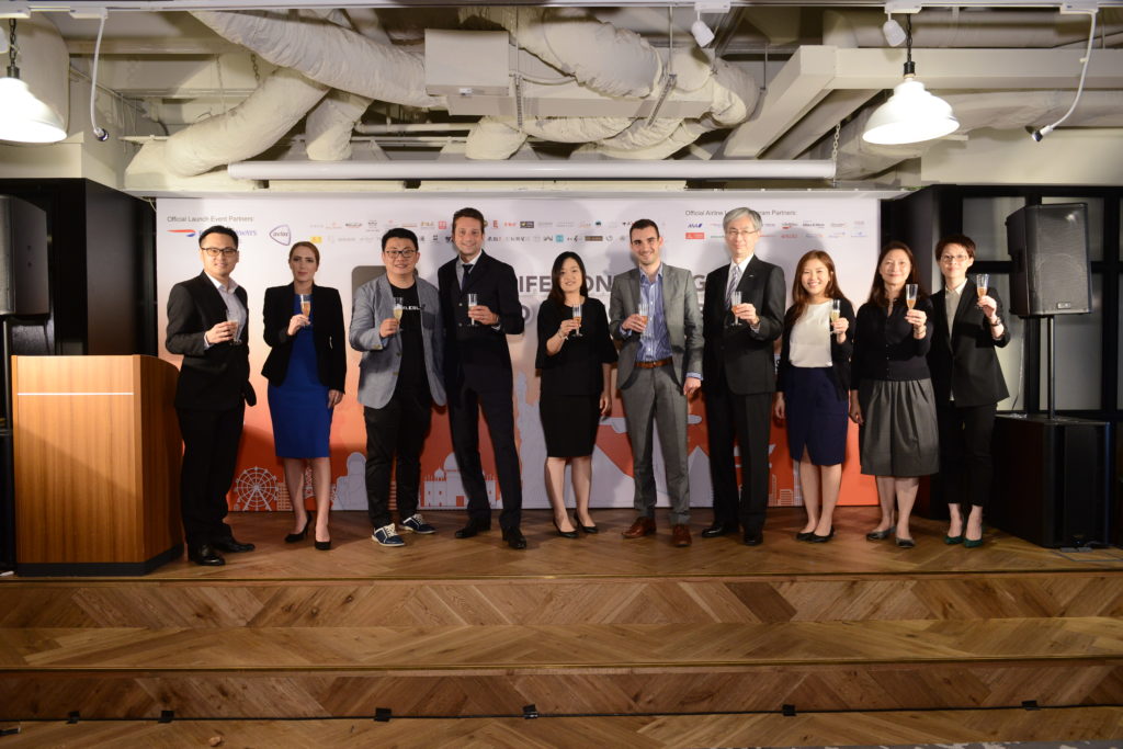 A group of ten people, dressed in business attire, are standing on a wooden stage in front of a backdrop with various logos. They are holding glasses and appear to be toasting or celebrating. The setting looks like an indoor event space with visible ceiling pipes and lighting fixtures. There is a podium on the left side of the stage.