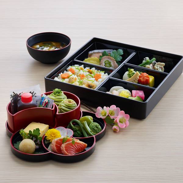 The image shows a beautifully arranged Japanese bento box meal. The bento box is divided into several compartments, each containing a variety of colorful and neatly arranged foods, including sushi, vegetables, and other traditional Japanese dishes. There is also a bowl of soup and a smaller, round container with additional items such as noodles, sauces, and garnishes. A small decorative flower is placed next to the bento box, adding to the aesthetic presentation.
