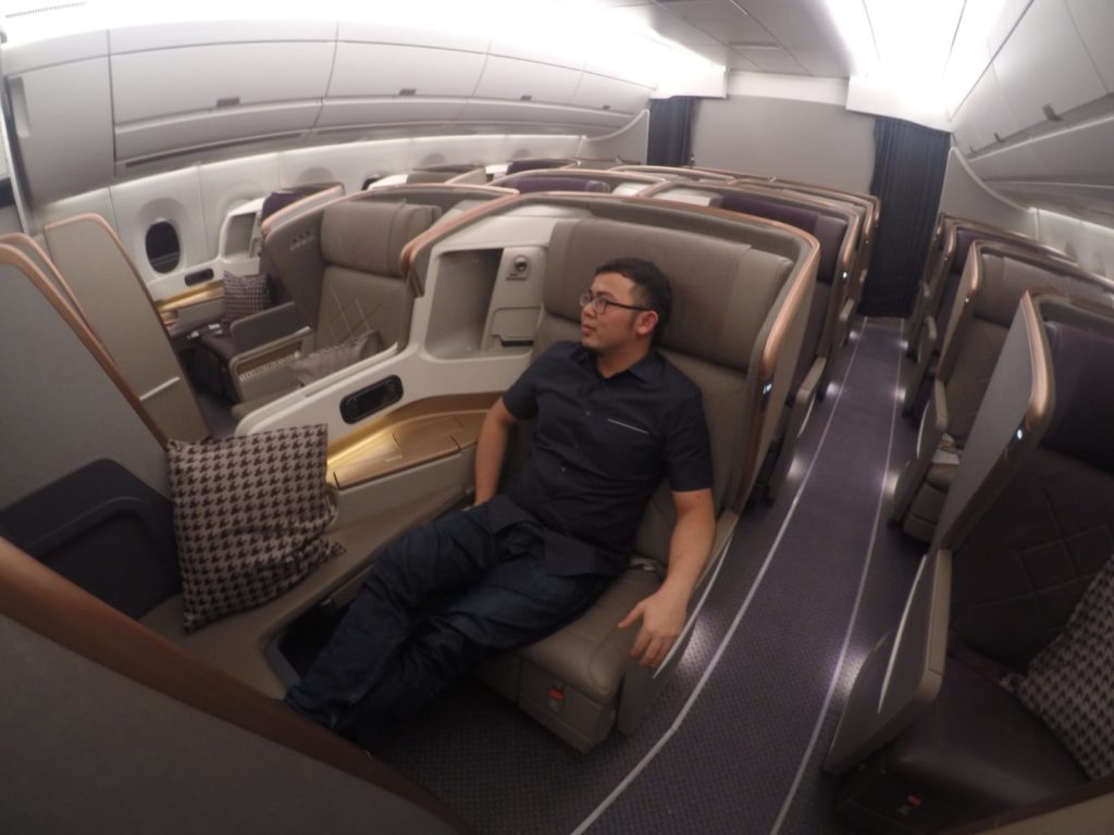 A man is seated in a luxurious first-class airplane cabin. The cabin features spacious, individual seats with high walls for privacy, plush upholstery, and ample legroom. The man is reclining comfortably, and there are pillows on some of the seats. The cabin has a modern design with soft lighting and a sophisticated color scheme.