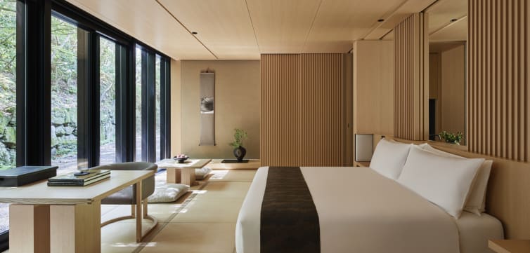 This image shows a minimalist, modern bedroom with a Japanese-inspired design. The room features a large bed with white linens and a dark throw blanket. There is a low wooden table with cushions on the floor for seating, and a small potted plant on the table. The room has large floor-to-ceiling windows on one side, allowing natural light to flood in. The walls and ceiling are made of light wood, and there is a decorative scroll hanging on the wall. The overall aesthetic is clean, serene, and uncluttered.