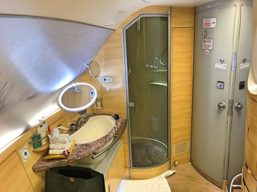The image shows the interior of a luxurious airplane bathroom. It features a modern sink with a marble countertop, a round illuminated mirror, and various toiletries neatly arranged. There is a glass-enclosed shower stall next to the sink. The walls and cabinetry are made of light-colored wood, giving the space a warm and elegant feel. The bathroom also includes a small towel on the floor and a black bag placed under the countertop. The overall design is sleek and sophisticated, indicating a high-end travel experience.